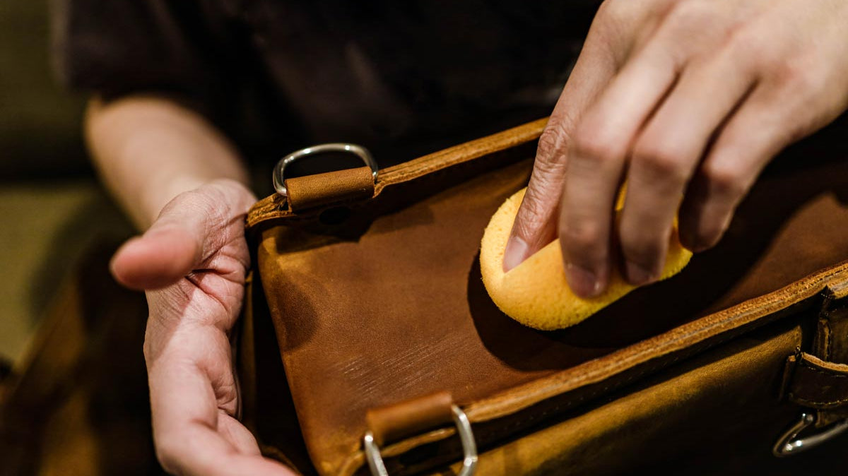 Closeup hand with yellow soft sponge cleaning stains on the surface of the vintage brown bag.